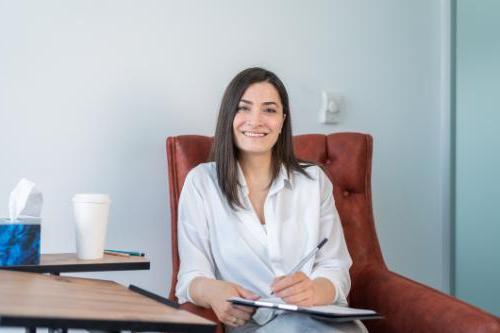 Smiling professional looking woman sitting in a chair in an office environment holding a clip board and pen.